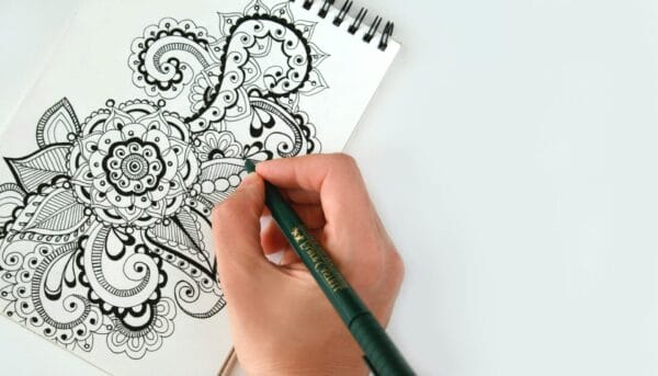 A hand is using a green pen to draw intricate black and white mandala patterns on a spiral-bound notebook, featuring floral and paisley designs against a white background.